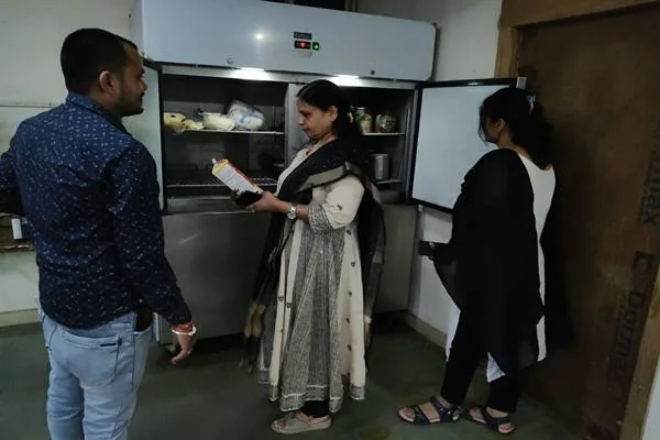 खुले रेफ्रिजरेटर के सामने खाद्य पैकेट देखते हुए महिला अधिकारी और पास खड़े दो लोग