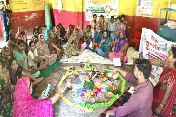 कमरे में पोषण सामग्री से सजी गोल प्रदर्शनी के चारों ओर बैठीं महिलाएं, किशोरियां और विभागीय प्रतिनिधि
