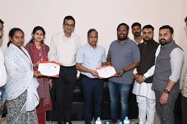 एक कमरे में खड़े महिला-पुरुषों के समूह के बीच दो लोगों को प्रमाणपत्र देते अधिकारी