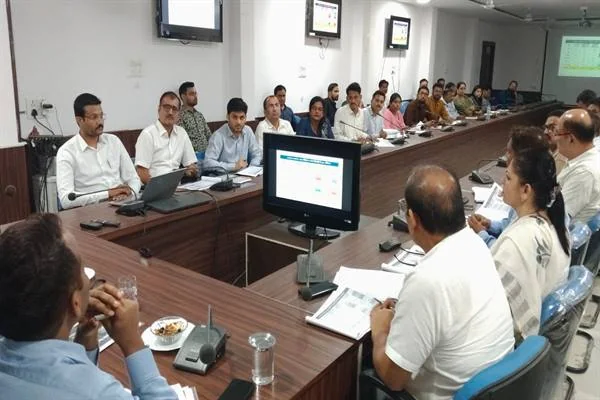 बैठक कक्ष में लंबी मेज के दोनों ओर बैठे अधिकारी, सामने कंप्यूटर स्क्रीन और दस्तावेज दिखाई दे रहे हैं।