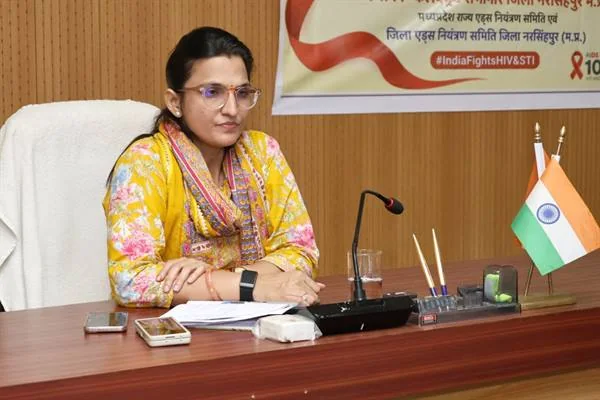 बैठक कक्ष में मेज के पीछे बैठी महिला अधिकारी, सामने माइक्रोफोन और दाईं ओर भारतीय ध्वज